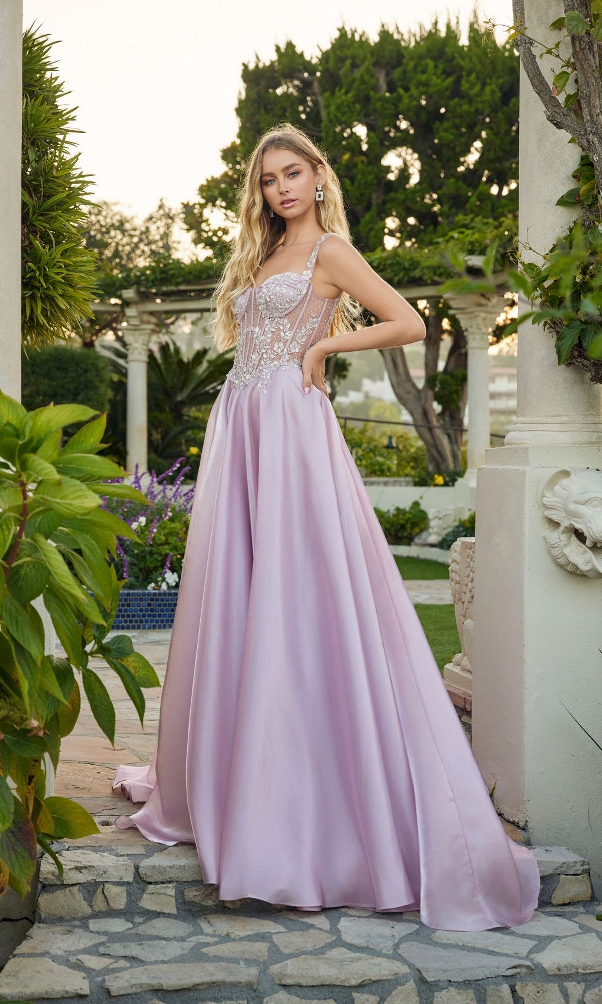 A woman poses outdoors in the Juliet JT2639G long prom dress in rose, featuring an a-line satin skirt and a sheer, beaded applique corset bodice. Stone columns and lush greenery appear in the background.